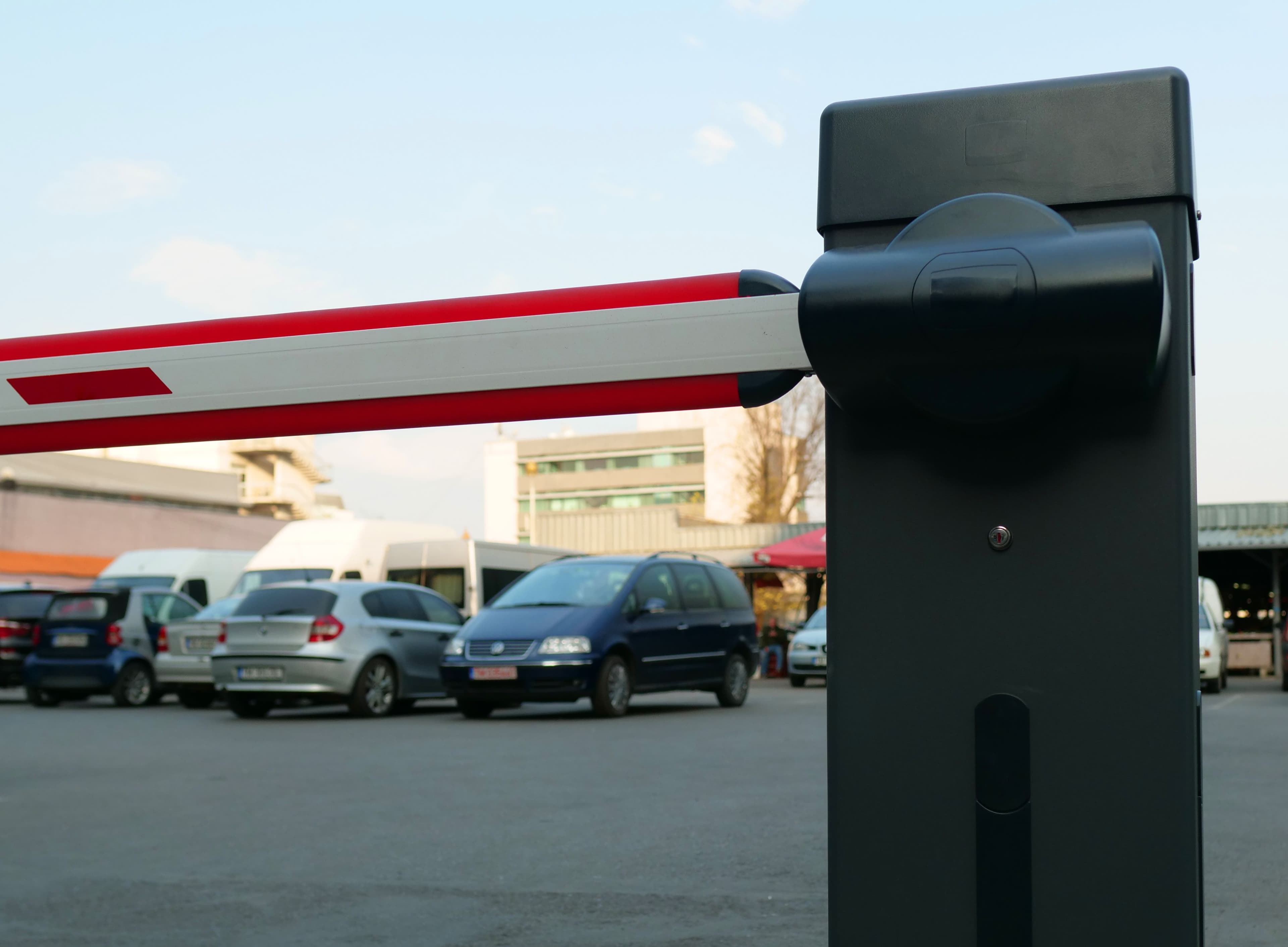 Red and white automatic barrier arm extended across the entrance of a busy parking lot.
